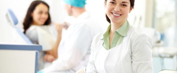 Pretty nurse looking at camera with smile on background of dentist and patient