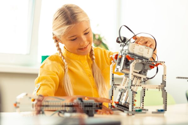 Cheerful girl sitting in the classroom playing with a robot she made at her school science lesson